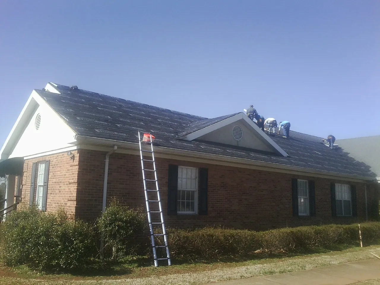 Crew tearing shingles off a brick ranch roof with a ladder set — Emergency Roof Repair work by our crew in Ocheyedan