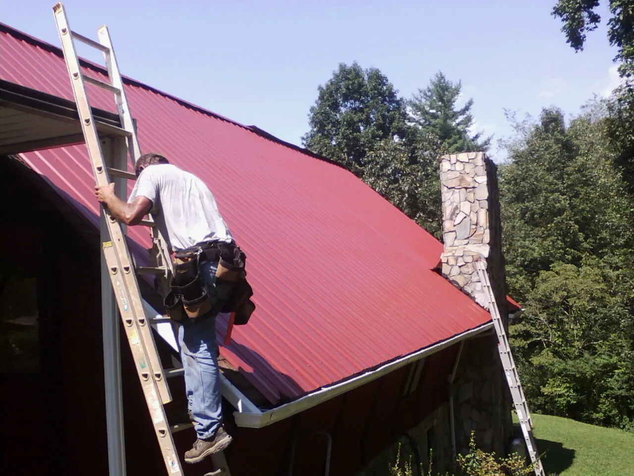 Roofer on a ladder beside a red metal roof and stone chimney — Soffit & Fascia Repair by Roofing Brothers in Ocheyedan